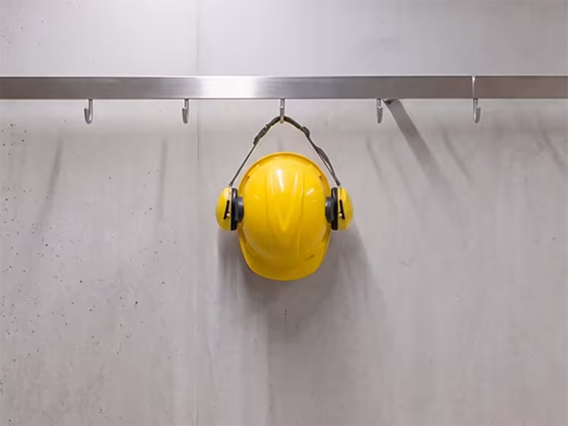 Casco de seguridad amarillo con orejeras colgando de un gancho en un perchero industrial, representando la eficiencia ante ventanas de parada muy cortas para mantenimiento.