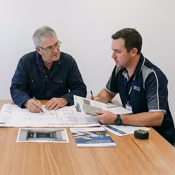 Dos profesionales de la construcción en una reunión de planificación, analizando planos arquitectónicos sobre una mesa de madera. Uno de ellos señala detalles en el plano mientras el otro sostiene una muestra de material y folletos corporativos.