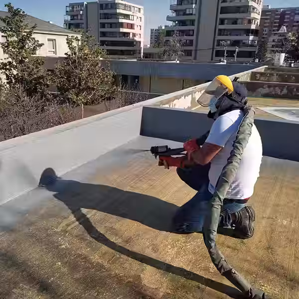 Operador con protector facial y EPP aplicando poliurea proyectada mediante pistola de alta presión sobre la losa expuesta de una azotea de hormigón en un edificio urbano.