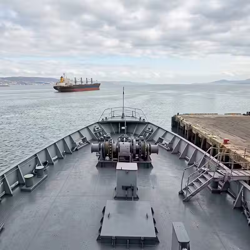Vista desde la proa de un buque mercante navegando cerca de un muelle, mostrando la superficie lisa de la cubierta de acero gris recién pintada y protegida.