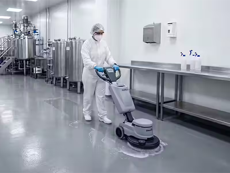 Industrias Una operaria con traje de protección blanco, mascarilla, gafas y cofia maneja una máquina fregadora industrial sobre un suelo gris brillante en una planta de producción limpia. Al fondo se observan tanques de acero inoxidable y mesas de trabajo relucientes.